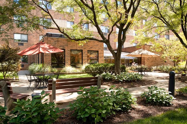 Outdoor courtyard area of a senior living facility with benches, tables with umbrellas, trees, and shrubs. The brick building of the facility is visible in the background with several windows.