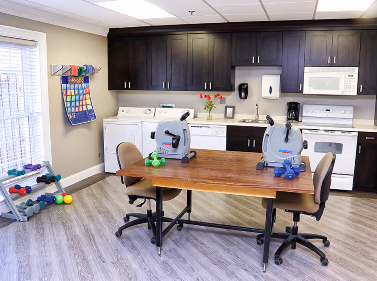 A room with a kitchen area featuring dark wood cabinets, a white stove, microwave, dishwasher, and washer and dryer. In front of the kitchen is a wooden table with two small exercise bikes and dumbbells on it. Two office chairs are positioned at the table. To the left, there is a rack with various dumbbells and colorful balls, and a calendar hanging on the wall near a window with blinds.