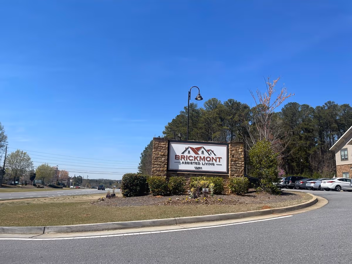 Entrance sign for Brickmont Assisted Living on a landscaped island with parked cars and trees under a clear blue sky.