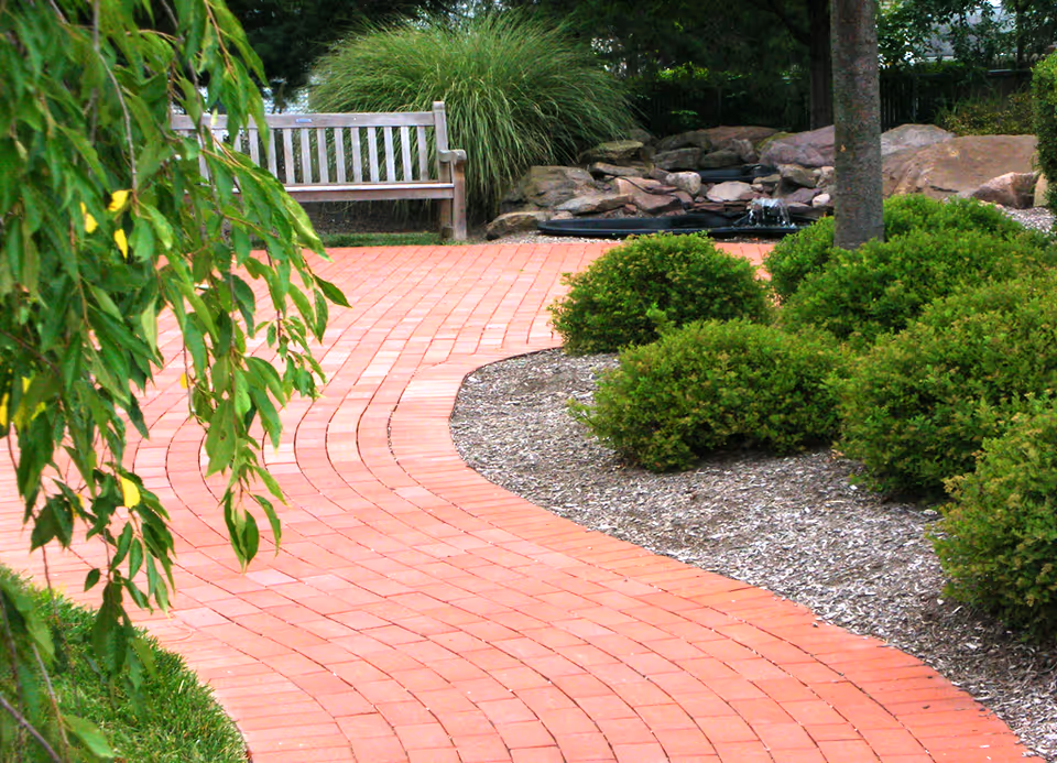 Curved red brick pathway in a garden area with green bushes, a tree, a wooden bench, and a small rock water feature in the background.