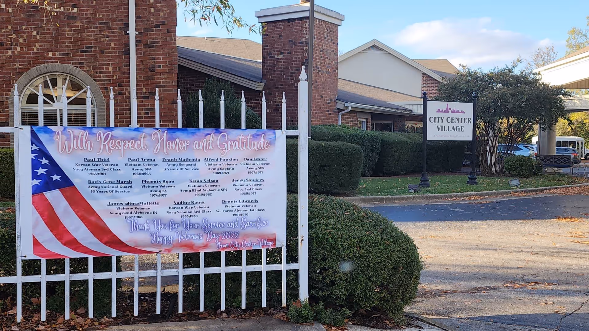 Entrance to City Center Village Senior Community with a white metal fence displaying a large banner honoring veterans. The banner features an American flag and lists names of veterans with a message of respect, honor, and gratitude for their service. Behind the fence is a brick building with a chimney and a sign for City Center Village near some bushes and trees.