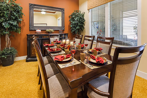 A warmly decorated dining room with a long wooden table set for a meal, chairs, a sideboard with a mirror, and potted plants.