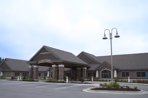 Front exterior of a single-story brick senior living facility with a covered entrance, stone columns, and a parking lot.