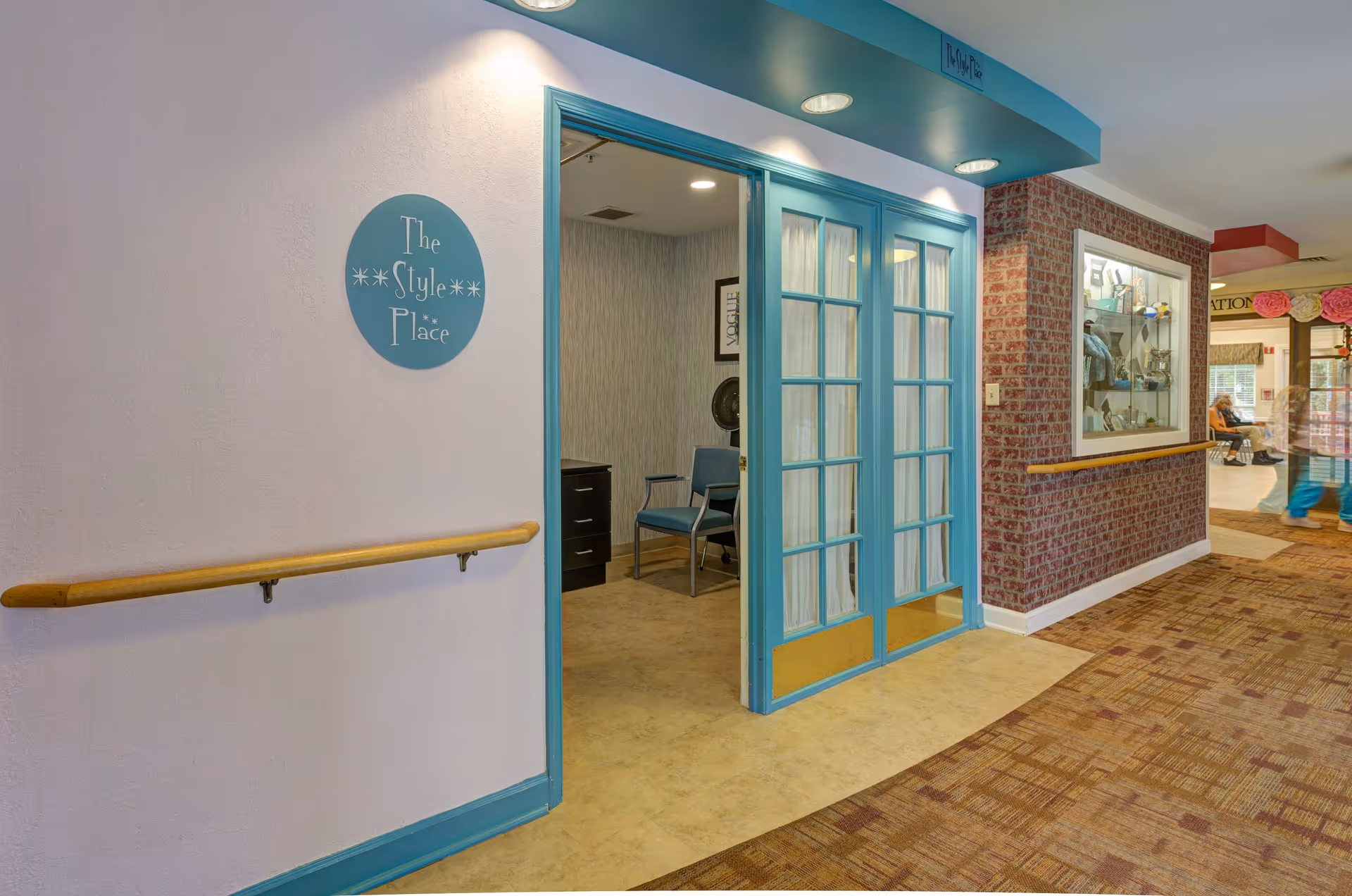 Interior hallway of a senior living facility with a blue-framed glass door entrance to a room labeled 'The Style Place'. The hallway has a wooden handrail on the left wall and a display case on the right wall. The floor is carpeted with a patterned design, and there is a person sitting in the background near another room.