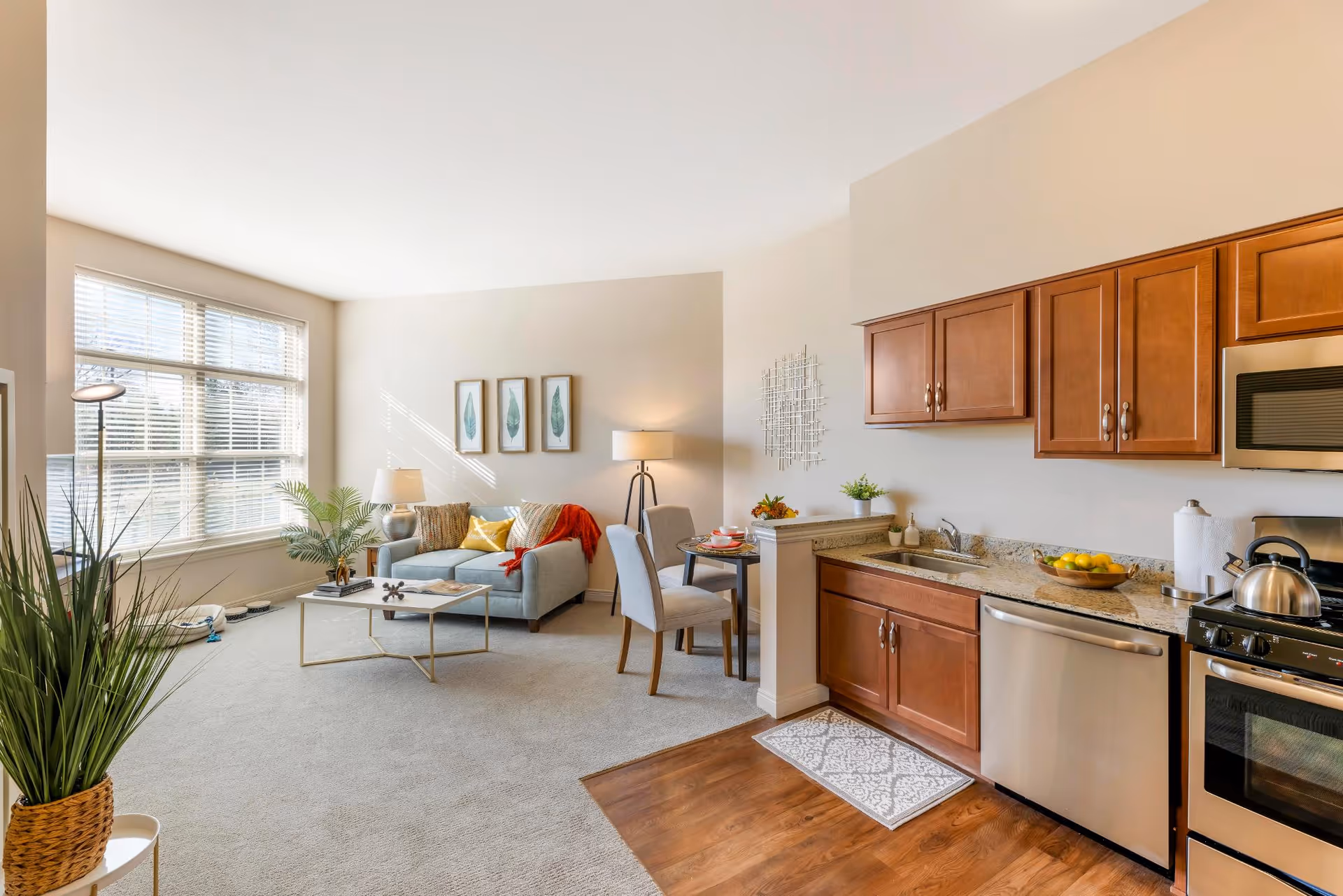 Bright and spacious living area with a large window letting in natural light. The room features a light gray sofa with colorful cushions and a red throw, a coffee table with decorative items, a small dining table with two chairs, and a kitchen area with wooden cabinets, granite countertops, and stainless steel appliances including a dishwasher and stove. There are plants and framed artwork on the walls, creating a cozy and inviting atmosphere.