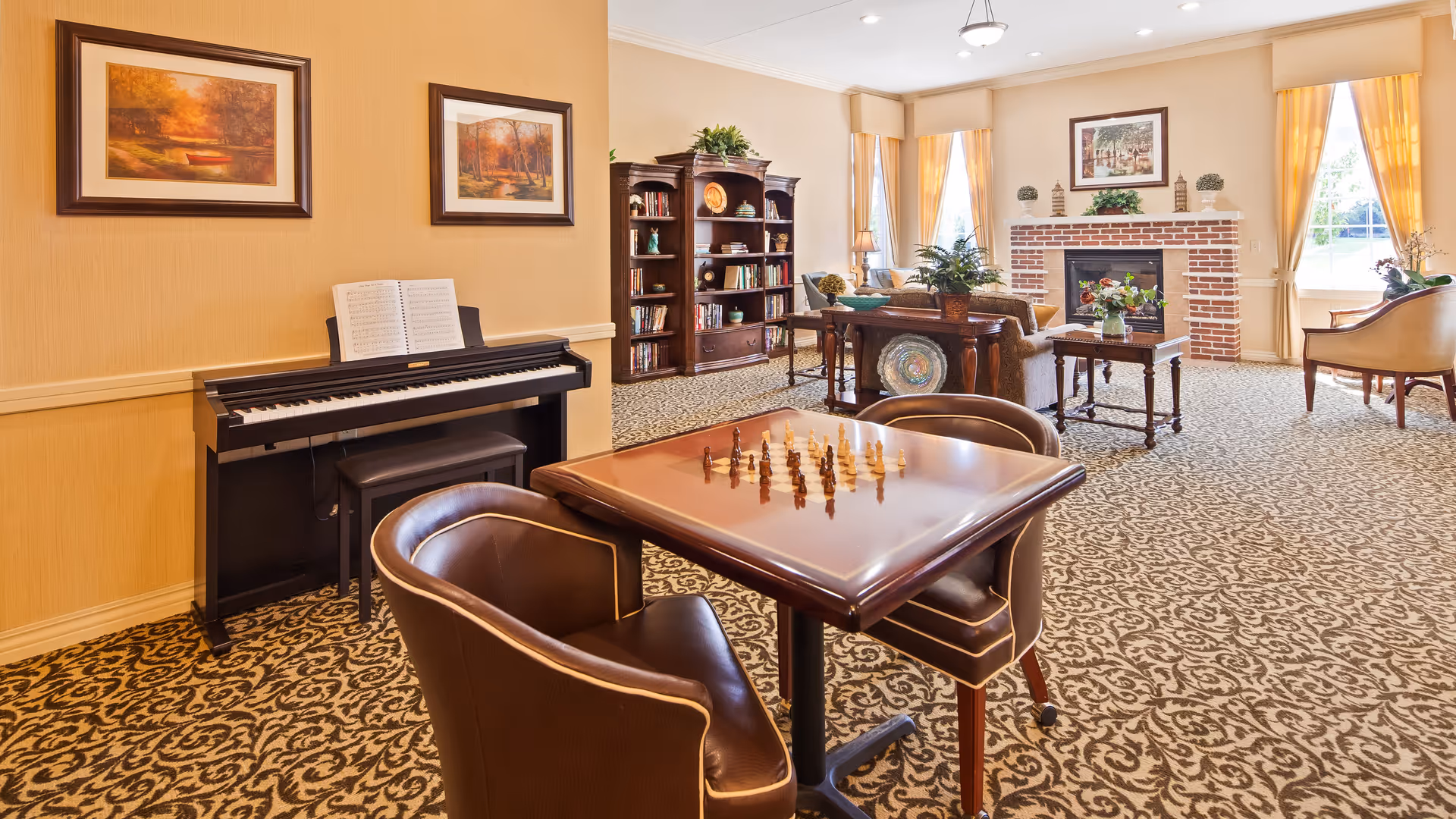 A spacious and well-lit common area with patterned carpet, featuring a chess table with two leather chairs in the foreground, an electric piano with sheet music against the wall, a wooden bookshelf filled with books and decorative items, a seating area with sofas and a coffee table, a brick fireplace, and large windows with yellow curtains letting in natural light.