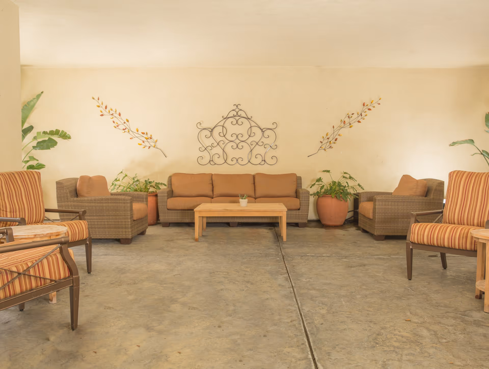 A cozy indoor seating area with a brown cushioned sofa and two matching armchairs arranged around a wooden coffee table. There are two striped cushioned chairs on either side, potted plants, and decorative wall art featuring metal scrollwork and leaf designs on a beige wall.