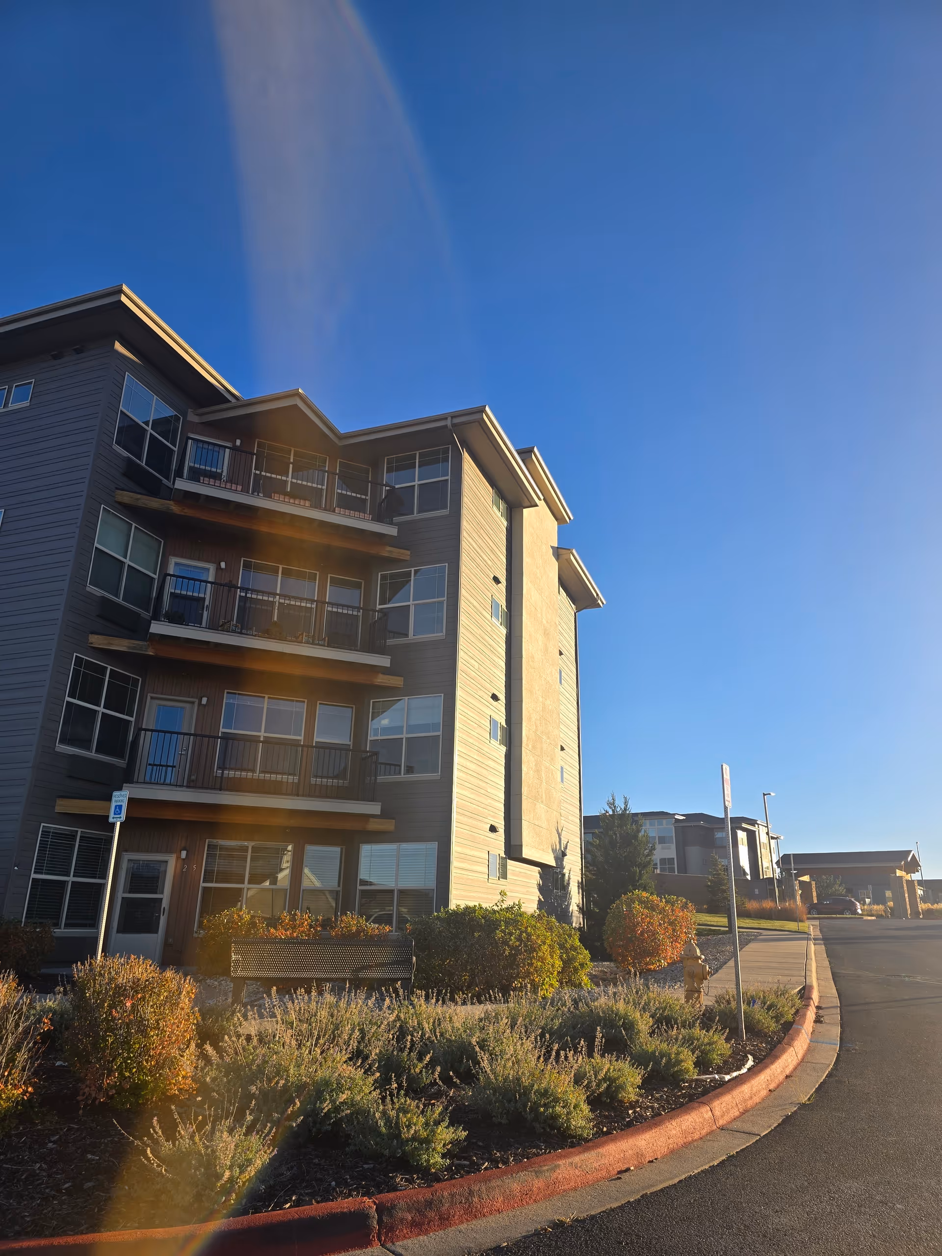 Exterior view of a multi-story residential building with balconies, surrounded by landscaped bushes and a bench near the sidewalk under a clear blue sky.