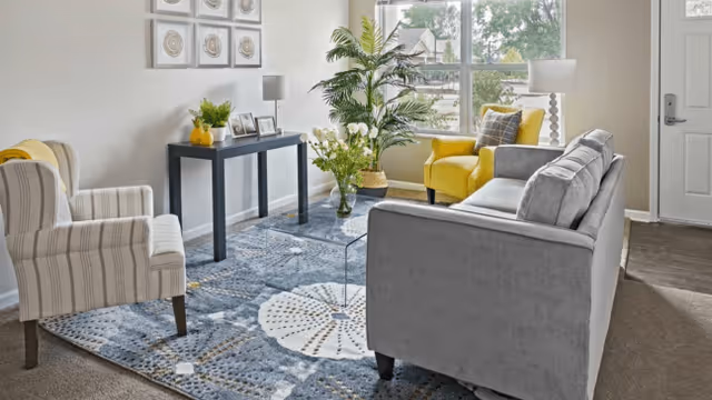 A bright living room with a gray sofa, a yellow armchair, and a striped armchair arranged around a glass coffee table on a patterned blue rug. There is a black console table against the wall with framed photos and a small plant. Large windows let in natural light, and a tall green plant is placed near the window. The room has neutral walls and a door visible on the right side.