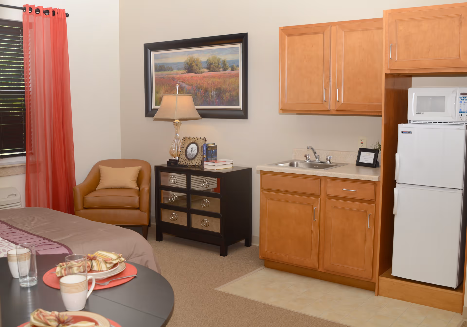 A cozy interior room featuring a small kitchenette with wooden cabinets, a sink, a microwave, and a refrigerator. To the left, there is a comfortable brown armchair with a beige pillow, a black dresser with mirrored drawers, a table lamp, and a framed landscape painting on the wall. In the foreground, a round table is set with plates, cups, and napkins. Red curtains cover the window on the left side.