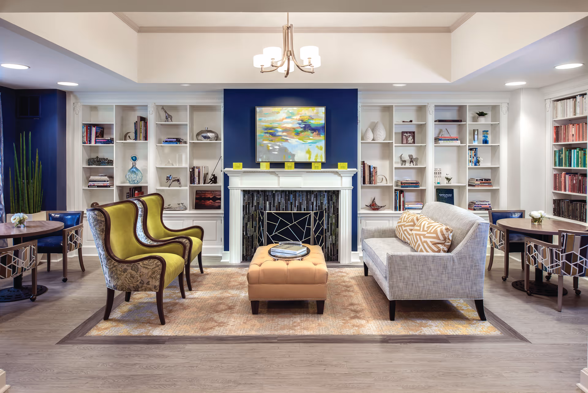 A cozy living room area in a senior living facility featuring a fireplace with a modern abstract painting above it. The room has built-in white bookshelves filled with books and decorative items on either side of the fireplace. There are two green upholstered armchairs with floral patterns, a gray sofa with patterned cushions, and a tan tufted ottoman in the center on a beige area rug. Two round wooden tables with chairs are placed on either side of the seating area. The room has a light wood floor and recessed ceiling lights.