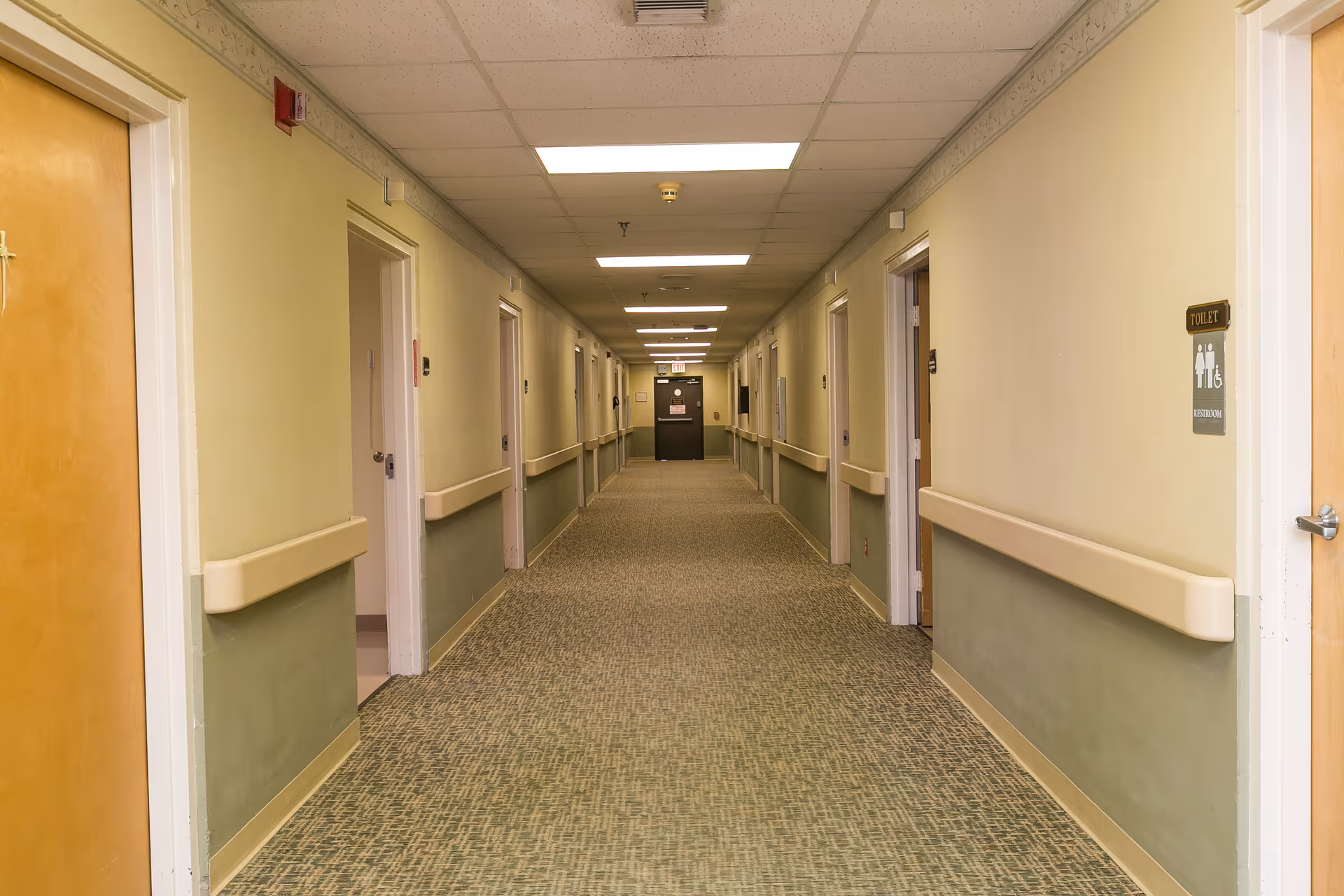 Long, well-lit interior hallway of a nursing facility with carpeted floor, handrails and doors to rooms on both sides.