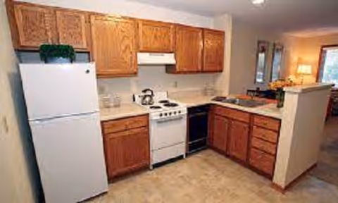 A kitchen area featuring wooden cabinets, a white refrigerator, a white stove with an oven, a black dishwasher, and a countertop with a sink. The kitchen opens into a living space with a lamp and window visible in the background.