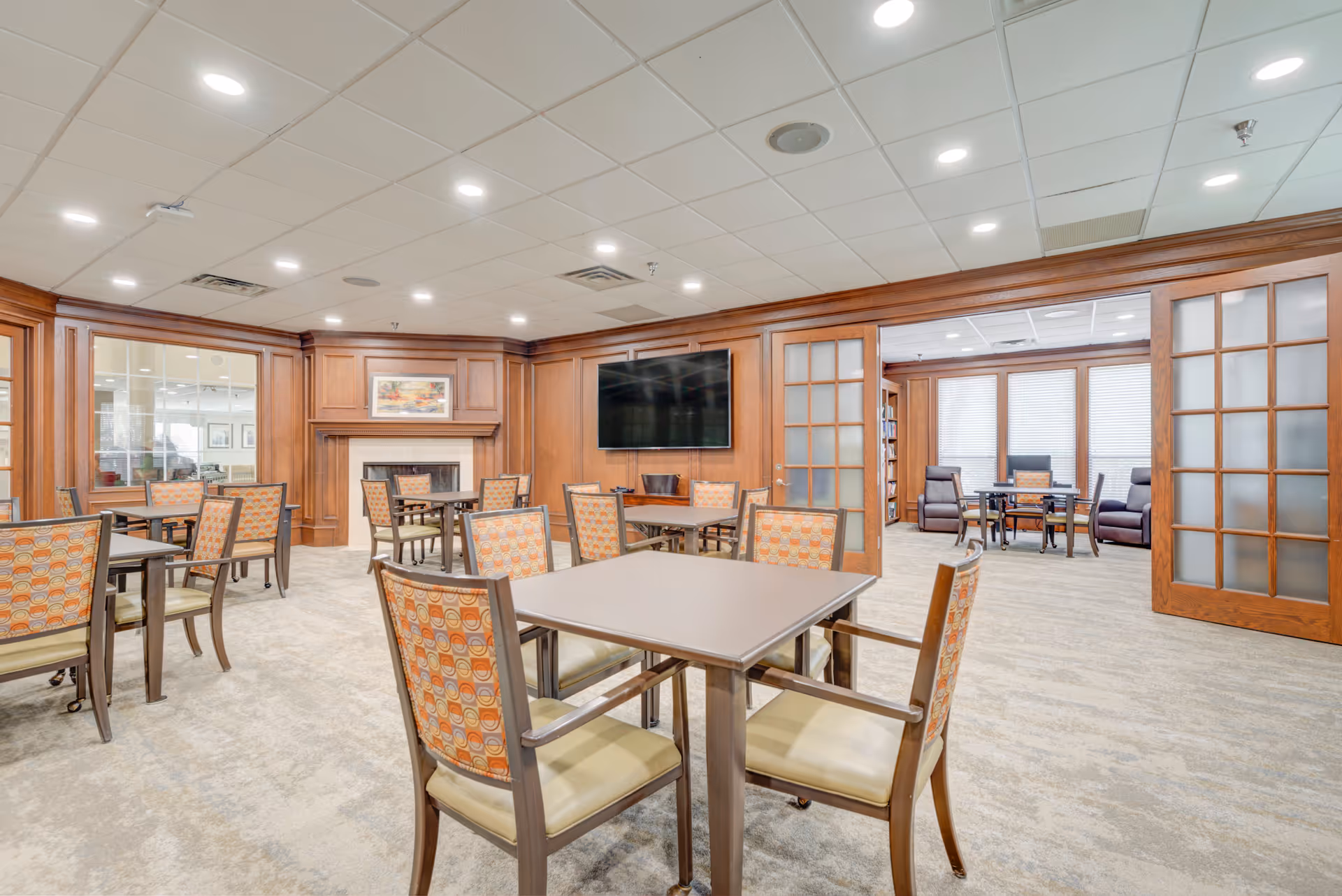 Bright communal dining/activity room with multiple tables and patterned chairs, a wall-mounted TV, fireplace, and a seating area visible through wooden glass doors.