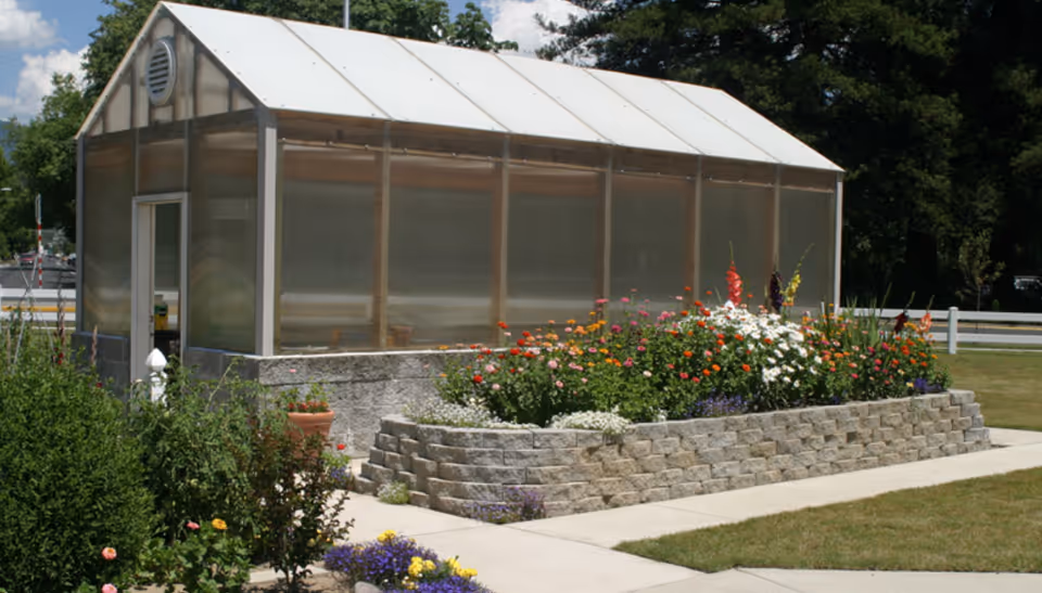 A greenhouse with translucent panels and a white roof situated next to a raised flower bed filled with colorful blooming flowers. The area is surrounded by a well-maintained lawn and a white fence in the background under a partly cloudy sky.