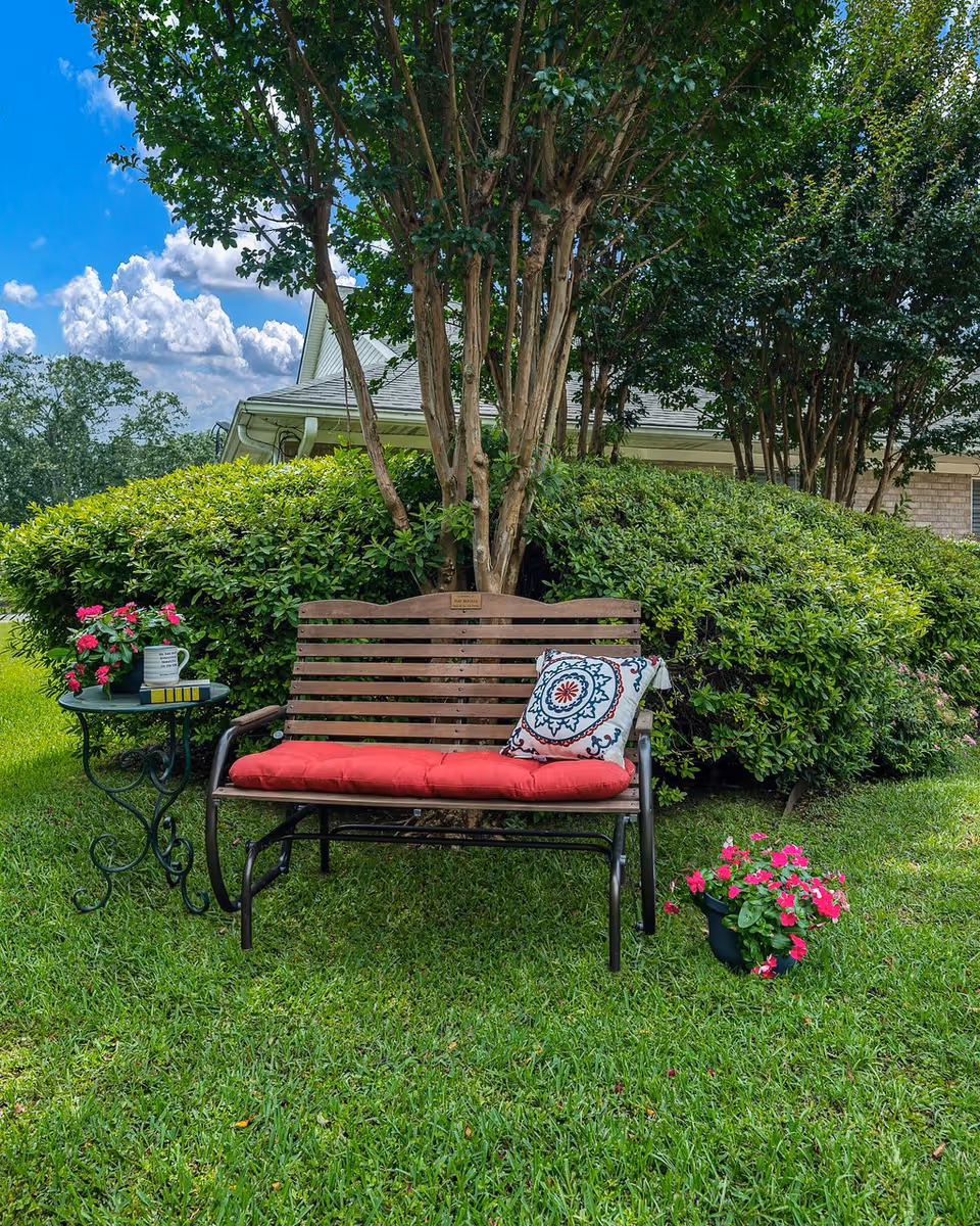 A wooden bench with a red cushion and a decorative pillow is placed on green grass in front of a large bush and tree. Next to the bench is a small round metal table with a potted pink flower and a white mug. Another pot of pink flowers is on the grass nearby. The sky is blue with scattered clouds, and part of a building roof is visible behind the greenery.