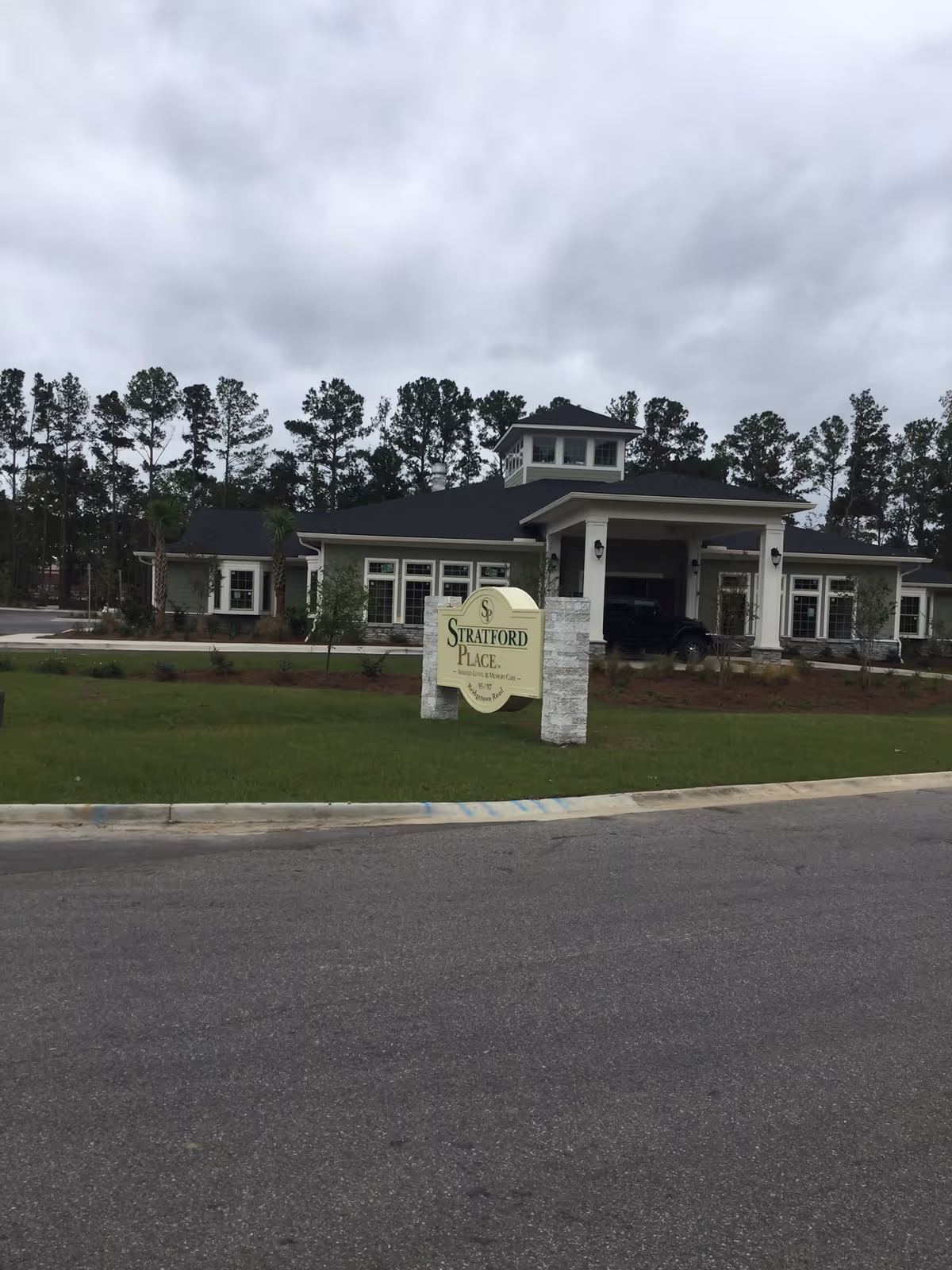 Exterior view of Stratford Place Assisted Living & Memory Care building with a sign in front on a grassy area. The building has a covered entrance and is surrounded by trees under a cloudy sky.