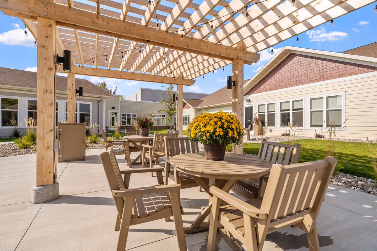Outdoor patio area at Fargo Elim Health Care Center with wooden pergola overhead, round tables with beige chairs, and potted yellow flowers on the tables. The patio is surrounded by a well-maintained lawn and buildings with beige siding and multiple windows under a clear blue sky.