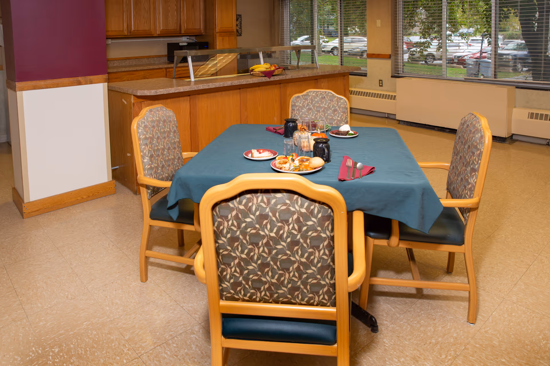 A dining area with a square table covered with a blue tablecloth, set with plates of food, utensils, and napkins. Four wooden chairs with patterned upholstery surround the table. In the background, there is a kitchen counter with a fruit bowl and large windows showing a parking lot outside.