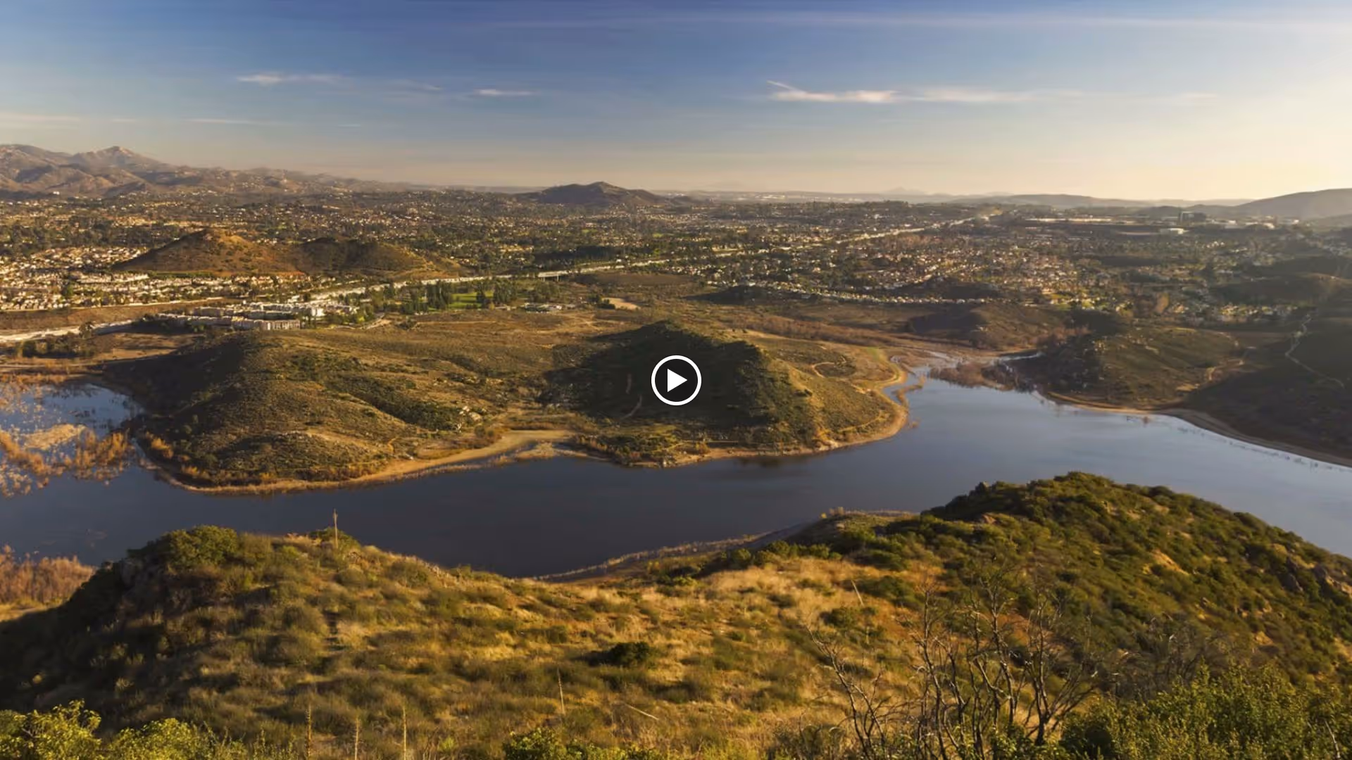 A panoramic view of a large body of water surrounded by hills and greenery, with a sprawling cityscape in the background under a clear sky during daylight.