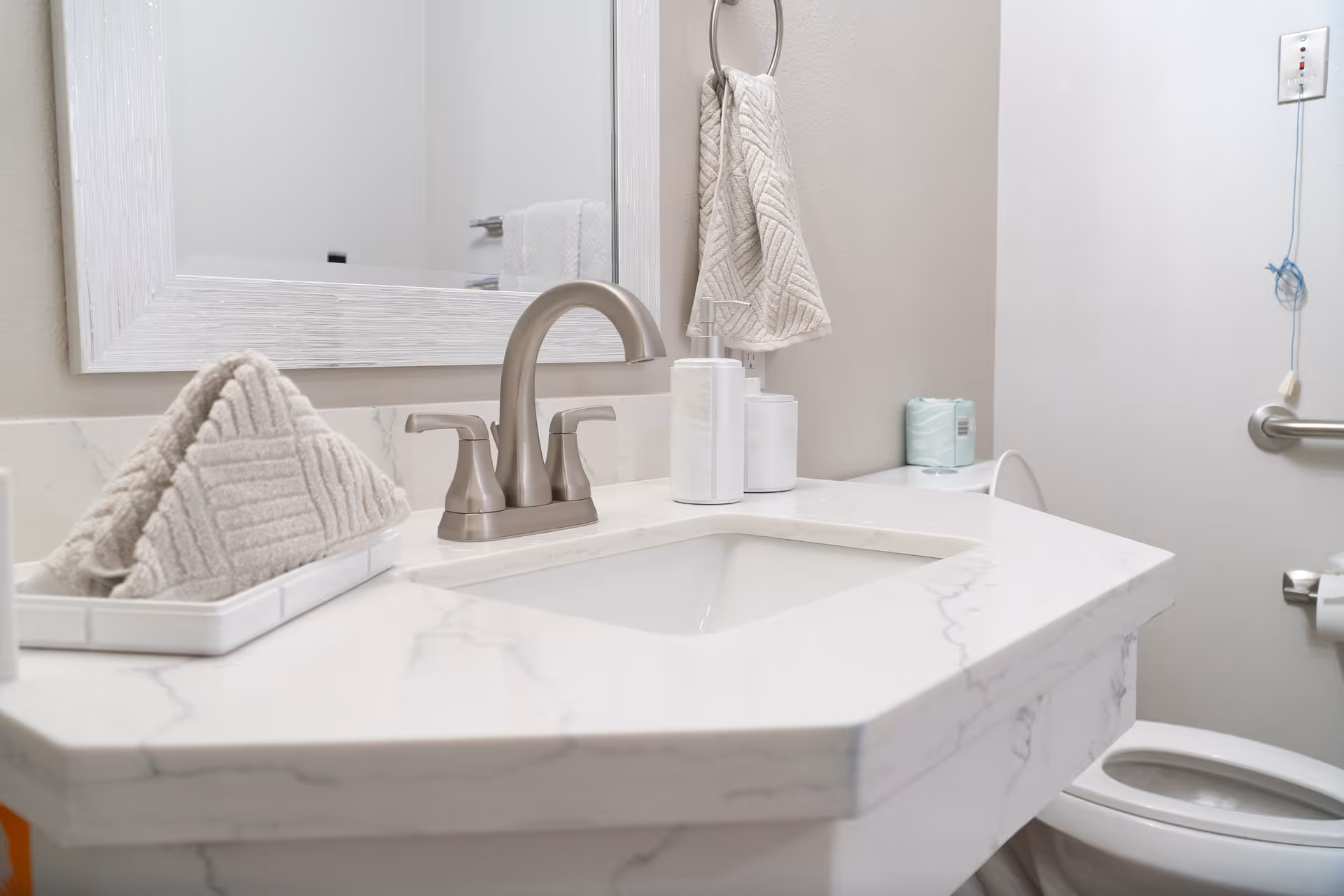 A clean bathroom sink area with a white marble countertop, a silver faucet, a large white-framed mirror, folded beige towels on a tray, a soap dispenser, and a hand towel hanging on a ring. A toilet with a grab bar and a toilet paper holder is visible to the right.