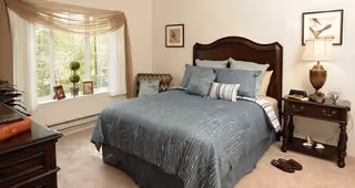 A cozy bedroom with a large bed covered in a blue-gray comforter and multiple pillows. There is a wooden nightstand with a lamp, decorative items, and a pair of slippers on the carpeted floor. A large window with sheer curtains lets in natural light, and framed artwork hangs on the walls.