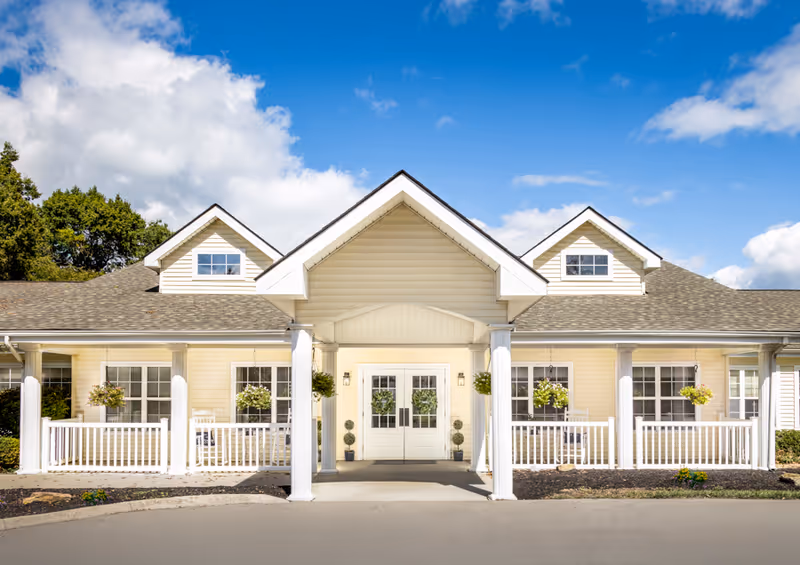 Front exterior view of a single-story building with a covered entrance supported by white columns, beige siding, multiple windows, and hanging flower baskets under a blue sky with some clouds.
