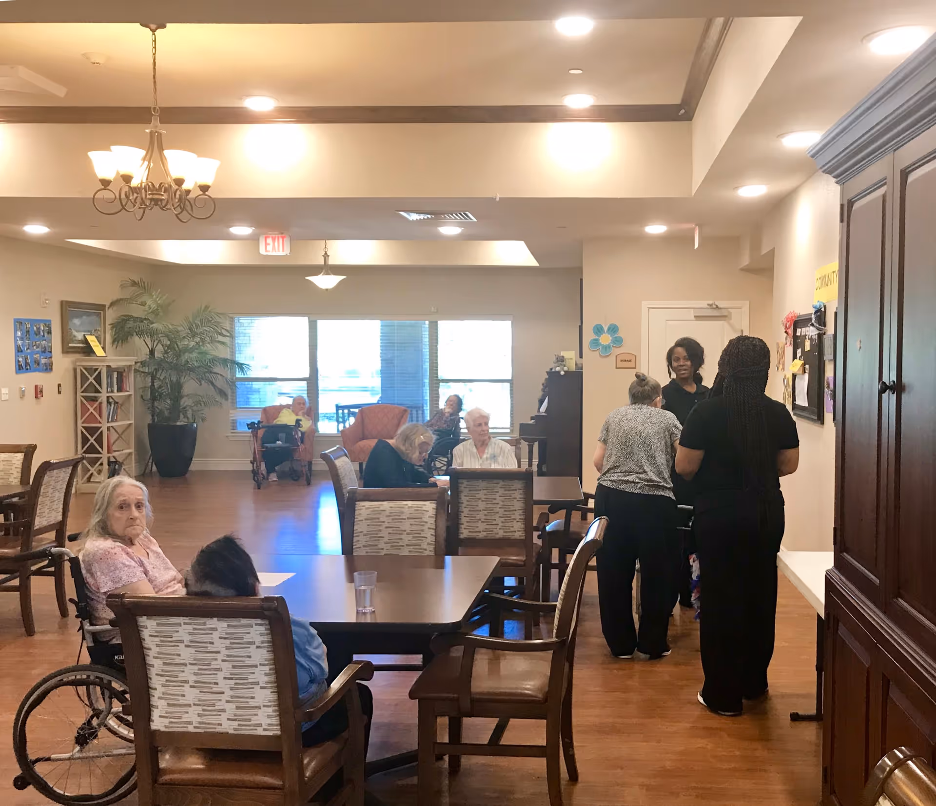 A common area in a senior living facility with several elderly residents seated around tables and in chairs. Some residents are in wheelchairs. Three staff members are standing and interacting near a bulletin board on the right side. The room is well-lit with ceiling lights and a chandelier, and there are large windows in the background letting in natural light. The floor is wooden, and there are plants and bookshelves along the walls.