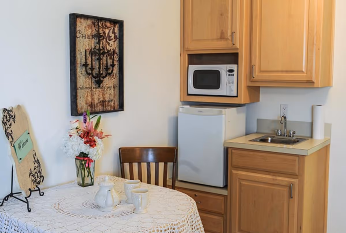 Small kitchenette and dining nook with a lace-covered table and floral centerpiece, wooden chair, mini refrigerator, microwave, sink, and cabinets.