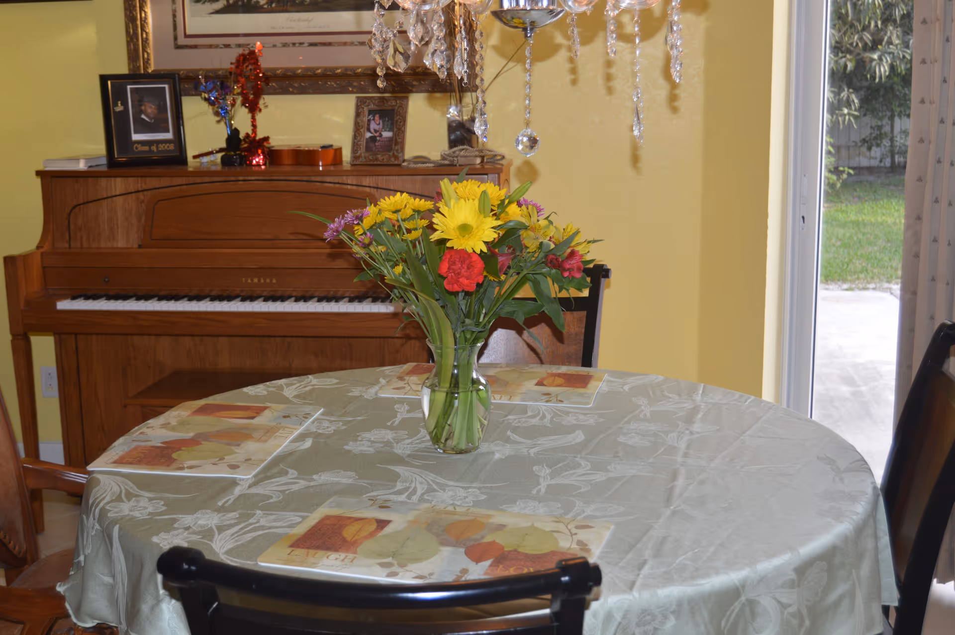 A round dining table covered with a light green tablecloth and decorated with a vase of colorful flowers in the center. Four placemats are set on the table. Behind the table is a wooden Yamaha piano with framed photos and decorative items on top. A crystal chandelier hangs above the table, and a sliding glass door with curtains is visible to the right, showing a glimpse of a green outdoor area.