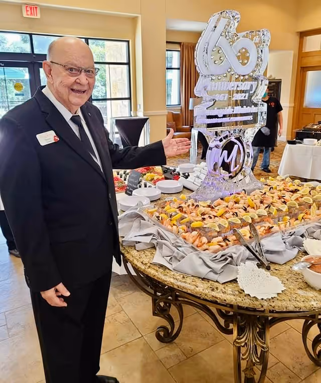 An elderly man in a black suit and tie stands indoors next to a large round table covered with a seafood spread and an elaborate ice sculpture. The man is smiling and gesturing towards the food and sculpture. The room has large windows and beige walls, with another person in the background near a buffet table.