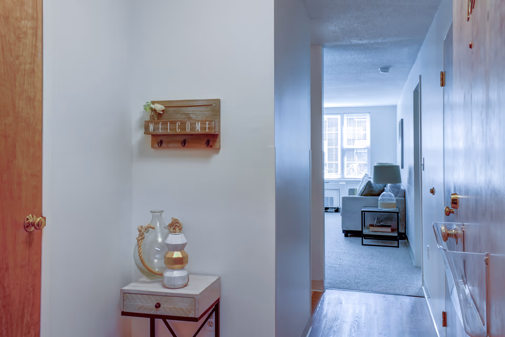 View down a hallway into a living room area with a gray sofa, side table with a lamp, and large windows letting in natural light. On the left wall of the hallway is a wooden 'WELCOME' sign with hooks and a small table below holding decorative vases. The hallway has light-colored walls and wooden doors.