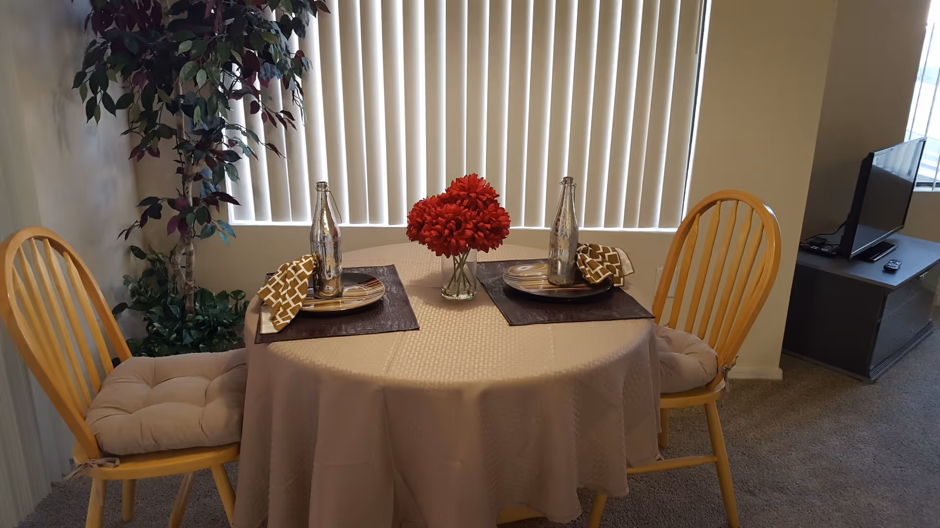 A small round dining table covered with a beige tablecloth set for two with plates, napkins, and decorative bottles. A vase with red flowers is in the center. Two wooden chairs with beige cushions are placed on either side of the table. A tall artificial plant is in the corner near vertical blinds covering a window. A TV on a stand is visible in the background.