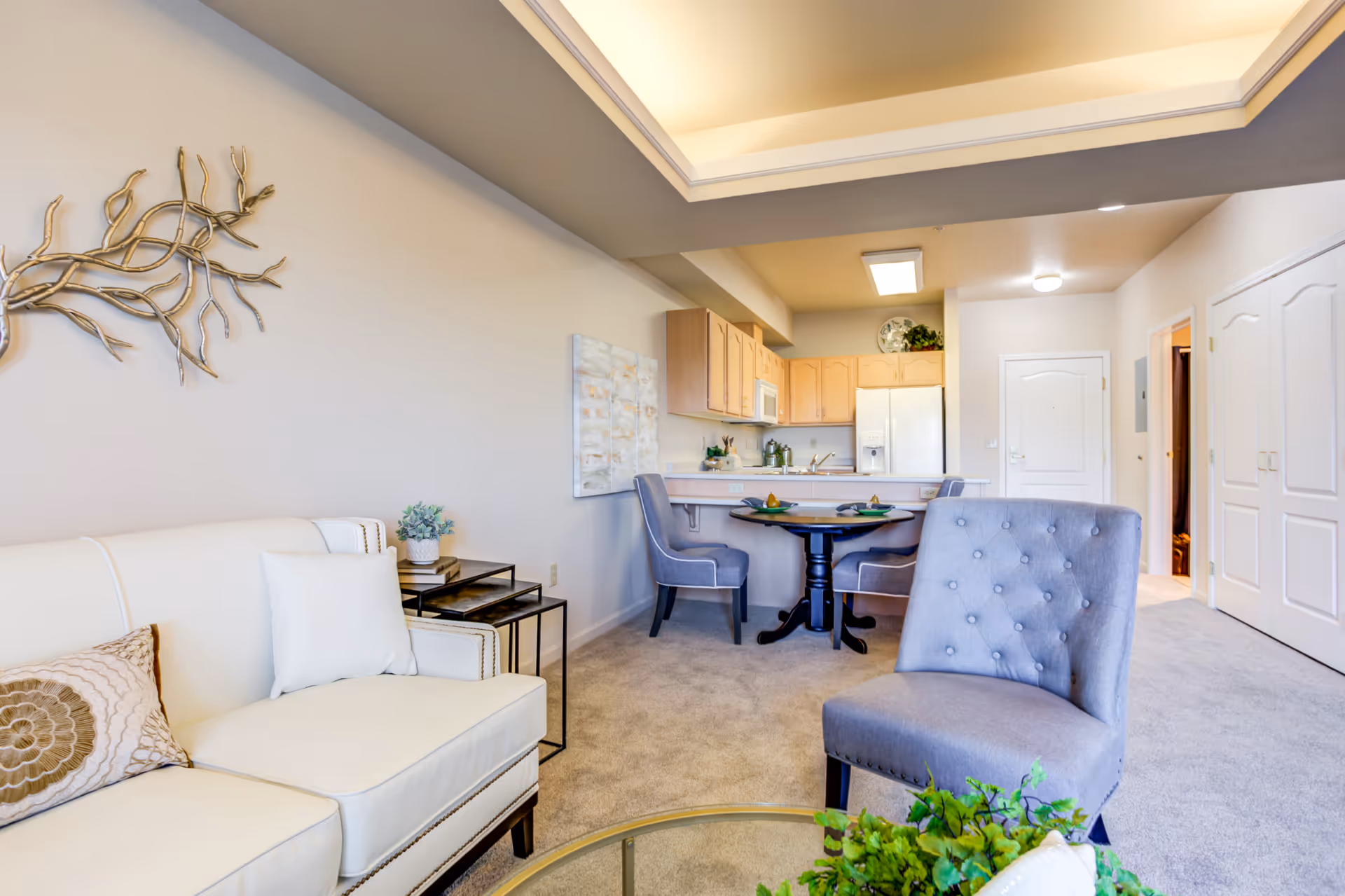 A bright and cozy living area in a senior living facility featuring a white couch with decorative pillows, a gray upholstered chair, a small round dining table with two chairs, and a kitchen area with light wood cabinets, a refrigerator, and a microwave. The room has beige carpet flooring, neutral-colored walls, and a decorative metal wall art piece.
