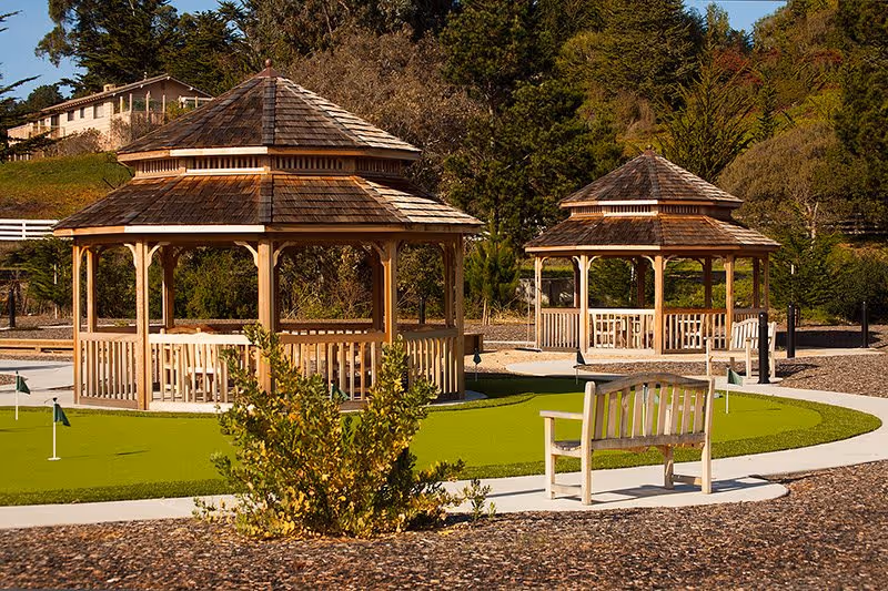 Two wooden gazebos with shingled roofs situated near a putting green with small flags, surrounded by trees and shrubbery under a clear sky.