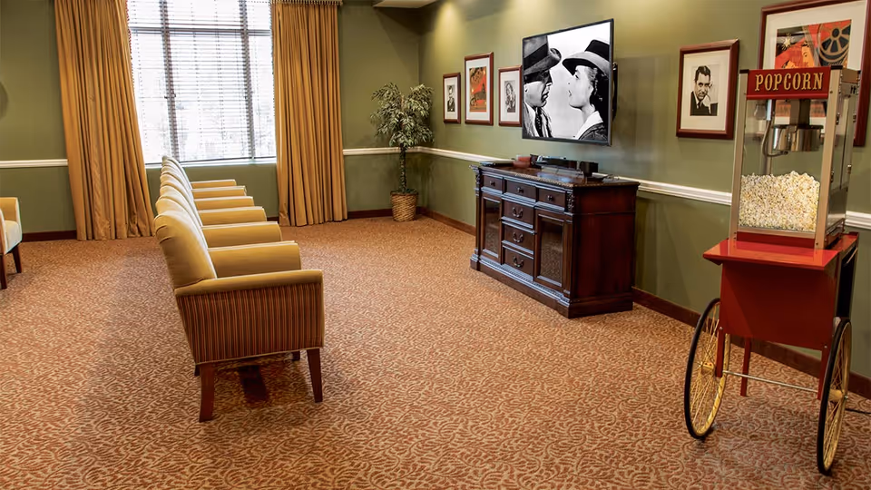 A cozy lounge area with a row of beige armchairs facing a large flat-screen TV mounted on a green wall. Below the TV is a dark wooden cabinet. The room has patterned carpet, a large window with beige curtains, framed black and white portraits on the wall, a potted plant in the corner, and a vintage-style popcorn machine on wheels.