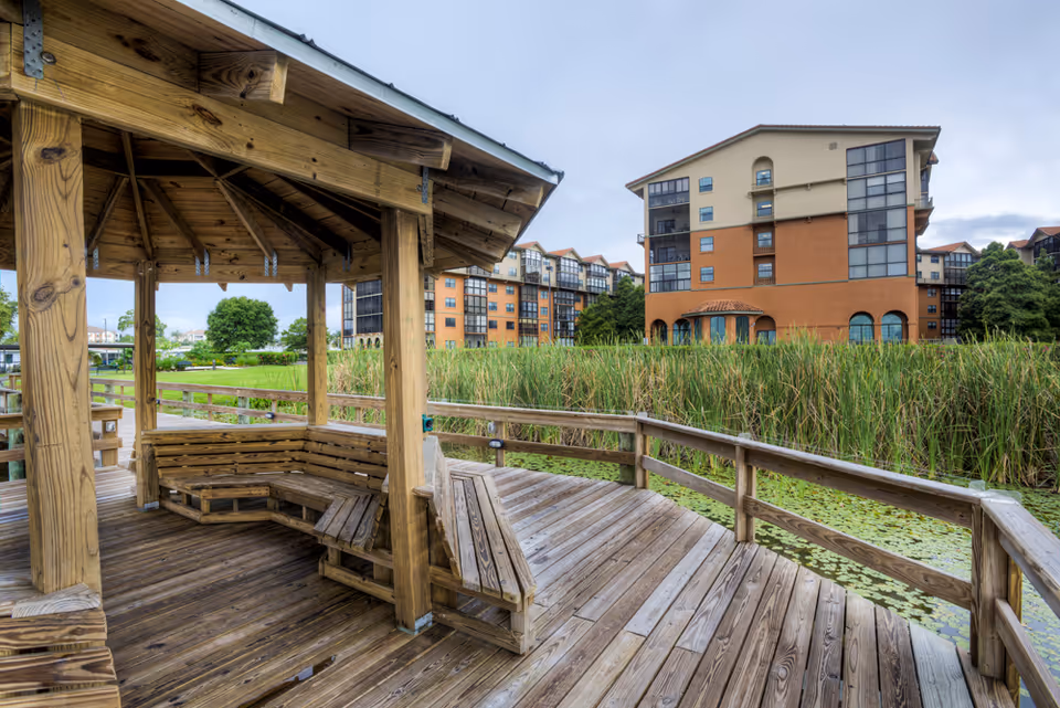 A wooden gazebo with built-in benches on a wooden deck overlooking a pond with lily pads and tall grass. In the background, there are multi-story residential buildings with a mix of beige and orange exterior walls under a cloudy sky.