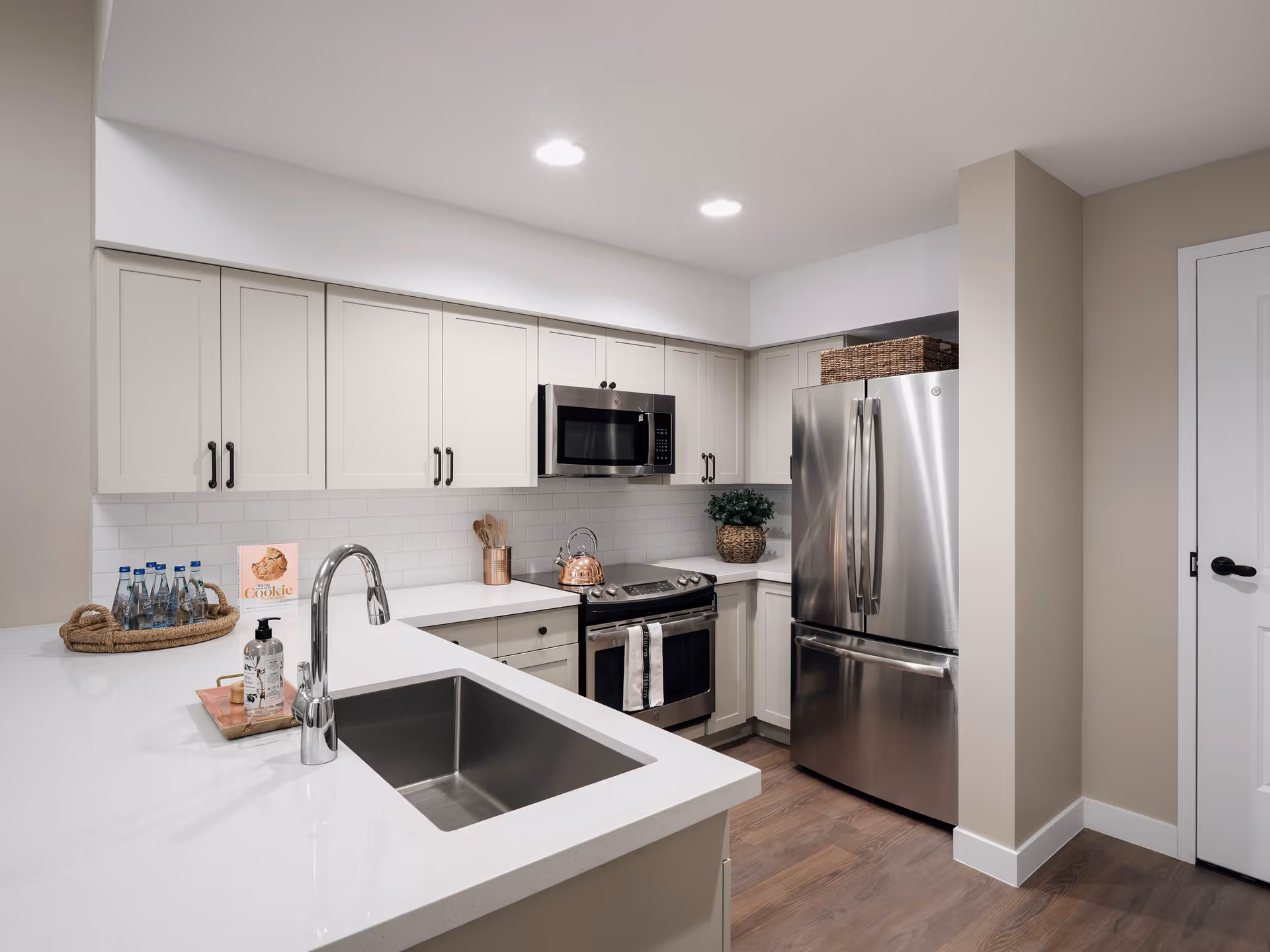 Modern kitchen with white cabinets, stainless steel refrigerator, microwave, and stove. The kitchen features a white countertop with a built-in sink and a chrome faucet. There are decorative items including a basket with water bottles, a small plant, and a cookbook on the counter.