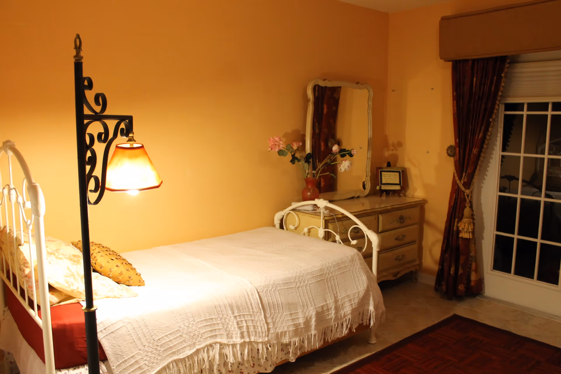 A cozy bedroom with a white metal bed frame, a white textured bedspread, and several pillows. There is a decorative floor lamp with a warm light next to the bed. Against the wall is a vintage-style dresser with a mirror, a vase with flowers, and a small framed item. The room has warm yellow walls, a window with dark red curtains, and a glass door leading outside.