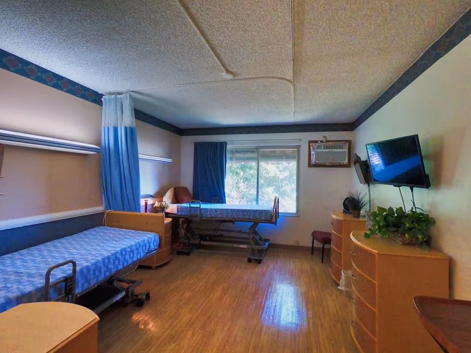 A room with two hospital-style beds separated by a blue privacy curtain. Each bed has a blue patterned mattress. There is a window with blue curtains letting in natural light. The room has wooden flooring, a wall-mounted TV, two wooden dressers with plants on top, and a small chair near the window.