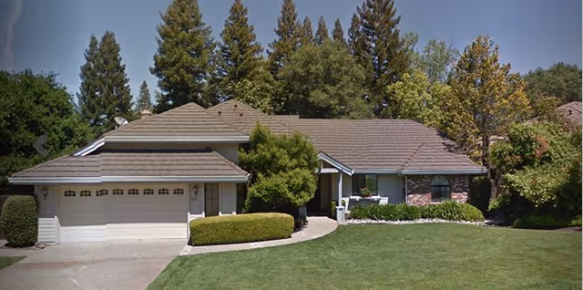 Single-story residential building with a two-car garage, surrounded by green trees and bushes under a clear blue sky.