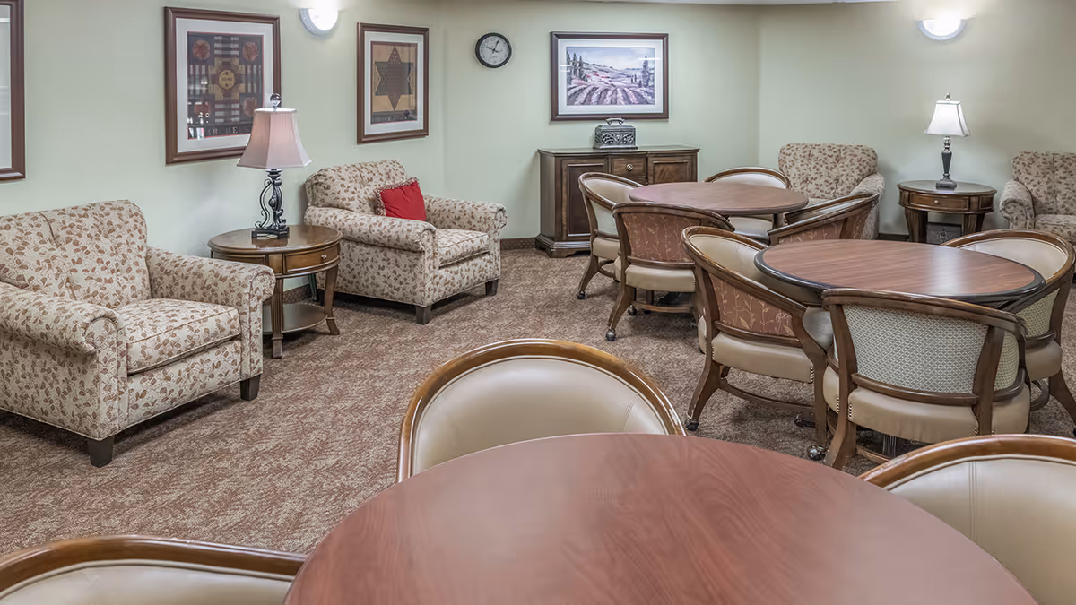 A senior living common room with round wooden tables, upholstered chairs, and patterned armchairs arranged on carpeted floor.