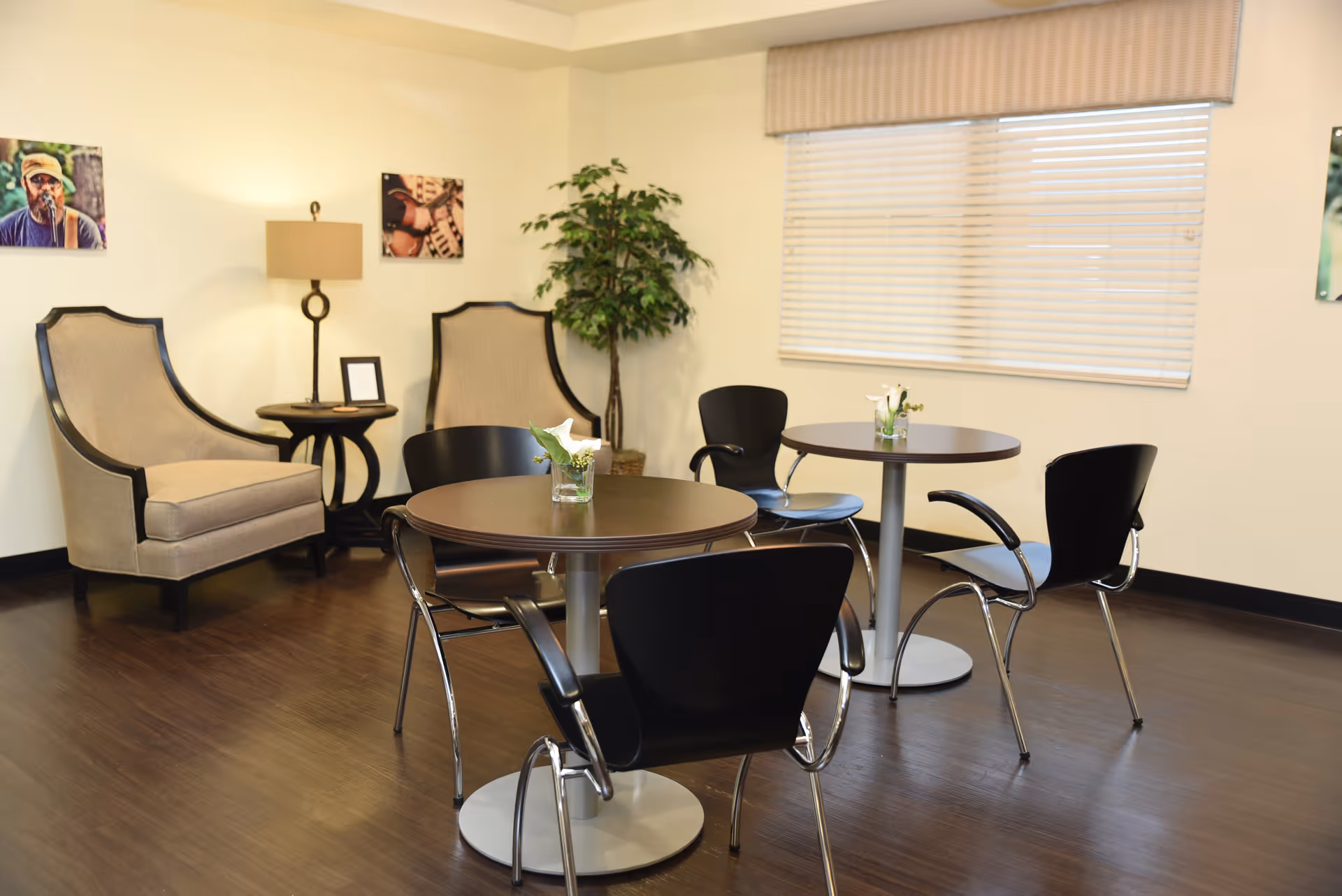 A cozy interior room with two round tables, each surrounded by three black chairs. There are two beige upholstered armchairs with dark wooden frames near a small round side table with a lamp. A potted plant is placed in the corner, and the walls are decorated with framed pictures. A window with closed blinds is visible on the right side.