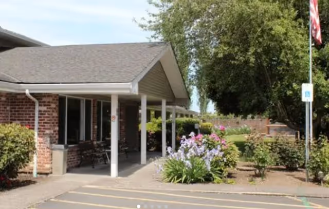 Exterior view of a single-story brick building with a covered porch supported by white columns. There are benches on the porch and a landscaped garden with flowering plants and bushes in front. A large tree and an American flag on a pole are visible near the garden. The scene is set on a clear day with a blue sky.