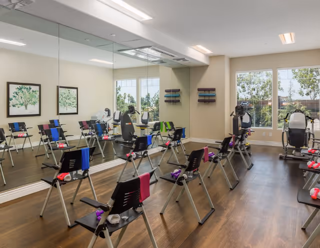 Bright exercise room with rows of folding chairs each holding small hand weights, large wall mirrors and cardio machines by windows.