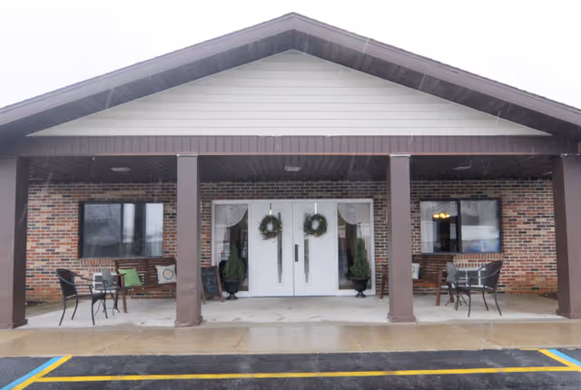 Front entrance of a brick building with a covered porch supported by four brown columns. The entrance features double white doors decorated with wreaths, flanked by two potted plants. There are two seating areas with chairs and small tables on either side of the entrance. The ground is wet, indicating recent rain.