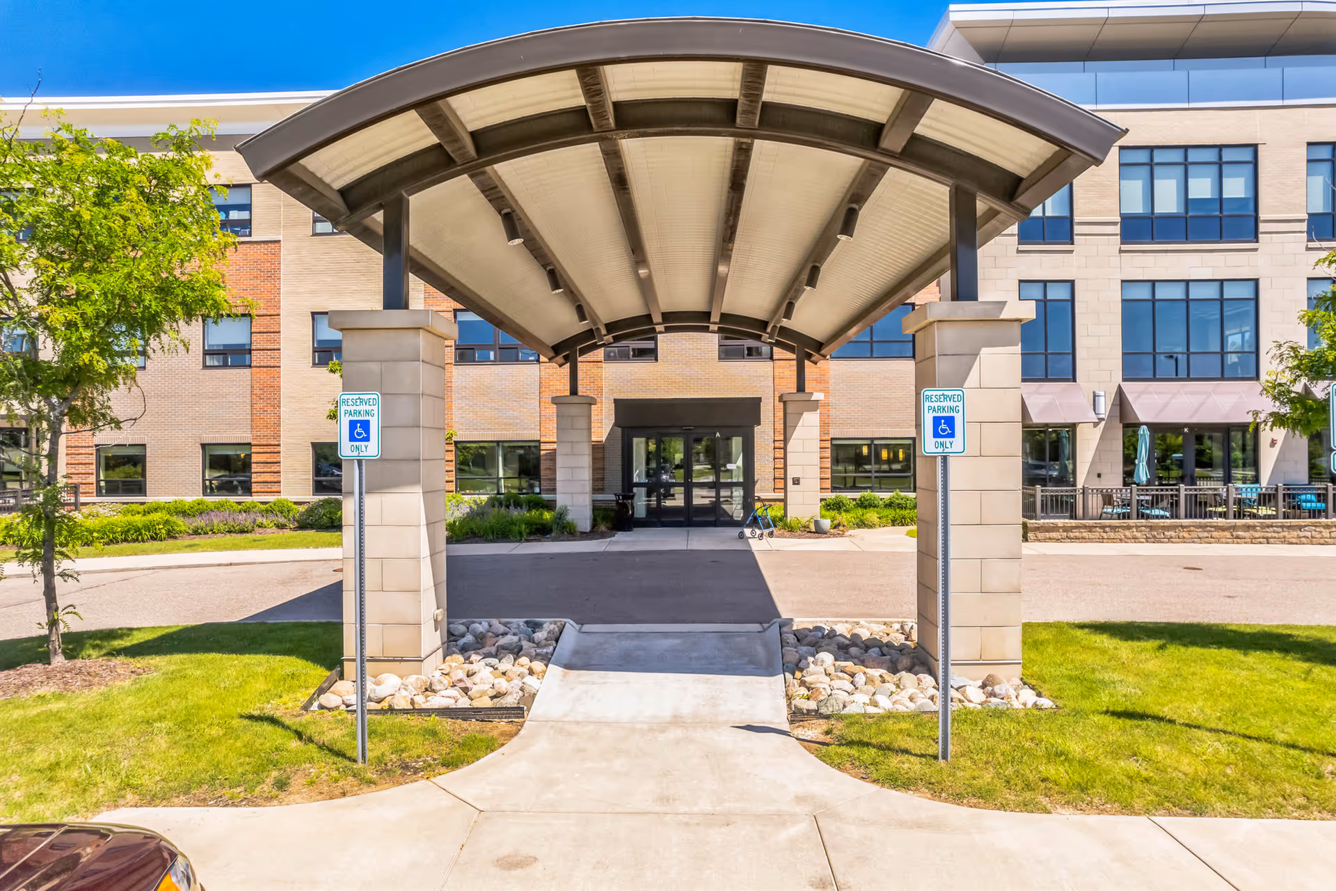 Entrance of a senior living facility named Willowbrook Hills with a covered drop-off area supported by stone pillars, reserved parking signs for handicapped access, and a multi-story building with large windows in the background under a clear blue sky.