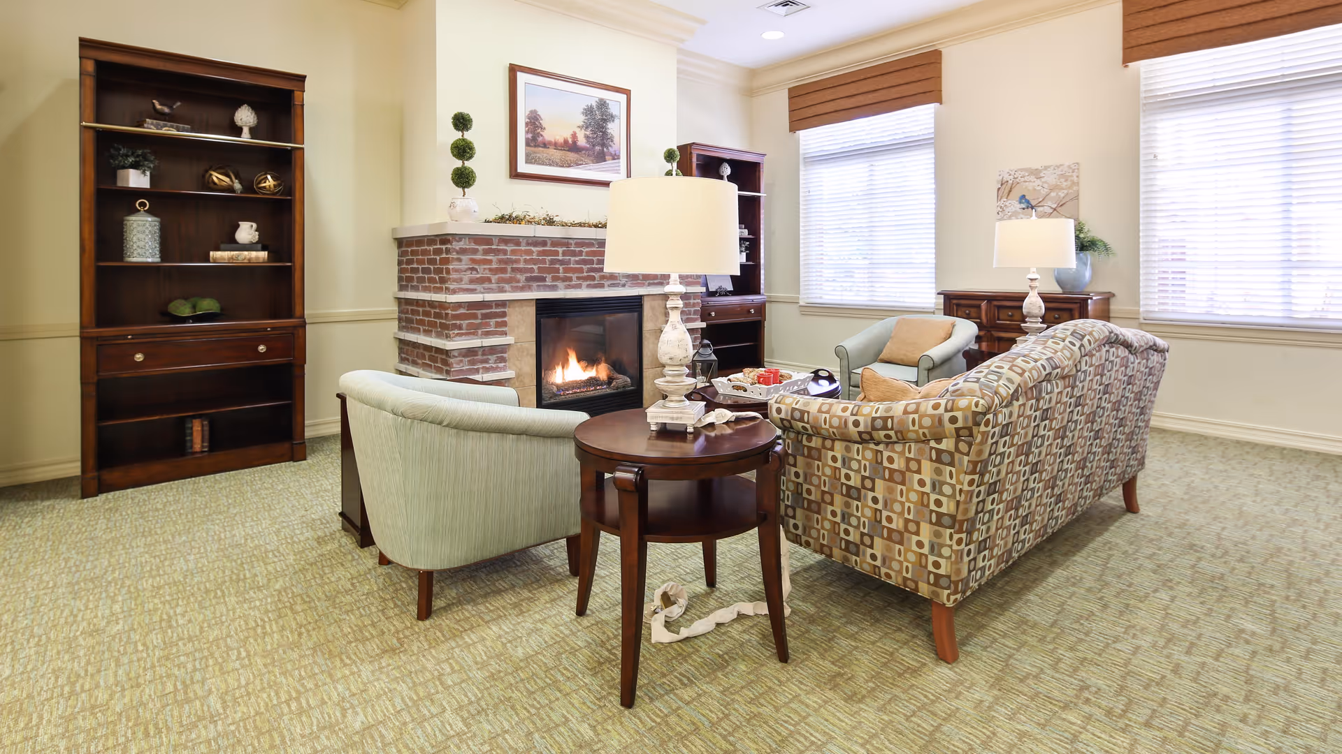 A cozy living room with a patterned sofa, two armchairs, a round wooden side table with a lamp, a brick fireplace with a fire burning, two wooden bookshelves, and two large windows with blinds letting in natural light.