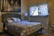 A cozy bedroom with a neatly made bed featuring a tufted headboard, patterned bedding, and multiple pillows. There is a nightstand with a lamp on one side of the bed, a window with sheer curtains allowing natural light to enter, and a large black and white cityscape photograph hanging above the bed.