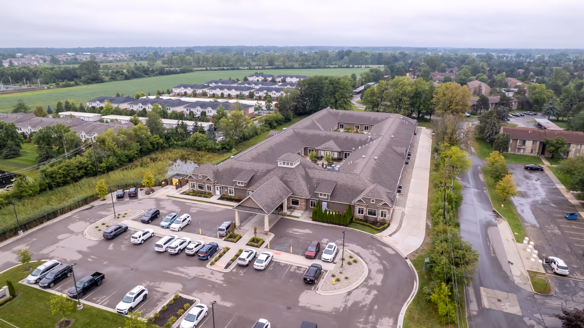 Aerial view of a large single-story senior living building with a covered main entrance, adjacent parking lot, and surrounding residential neighborhood and fields.